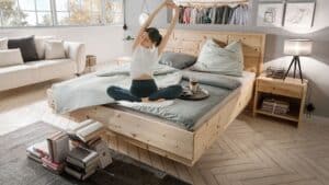 Helles Schlafzimmer mit Massivholzbett (z. B. Zirbe), ruhigen Farben und Leselampe; Person streckt sich morgens – Einrichtung, die Schlaf verbessern kann.