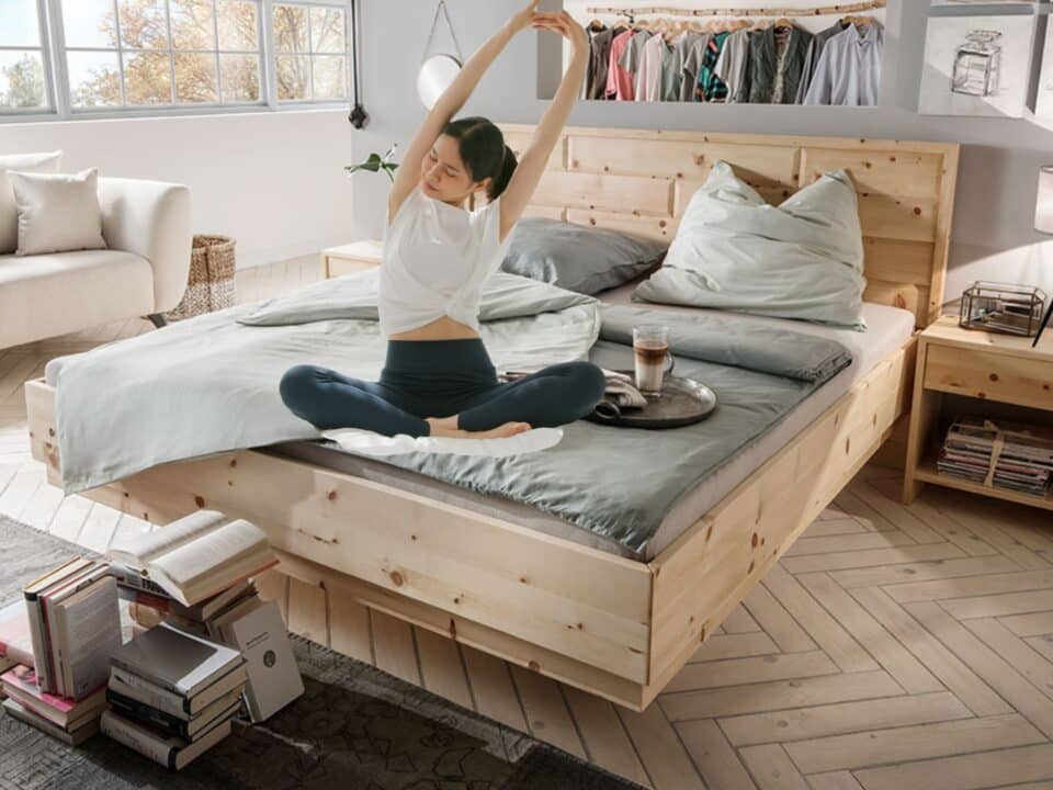 Helles Schlafzimmer mit Massivholzbett (z. B. Zirbe), ruhigen Farben und Leselampe; Person streckt sich morgens – Einrichtung, die Schlaf verbessern kann.